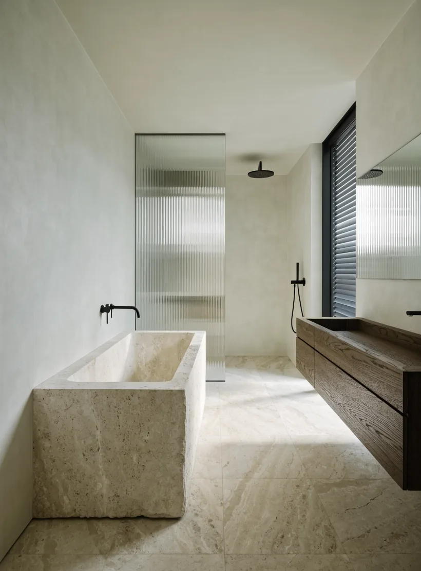 A bathroom with a bath carved from one block of travertine, a fluted glass shower screen, black tapware and a floating dark oak vanity