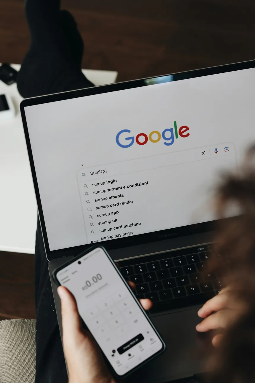 A hand holding a phone beside a laptop with a Google search box open and suggestions showing