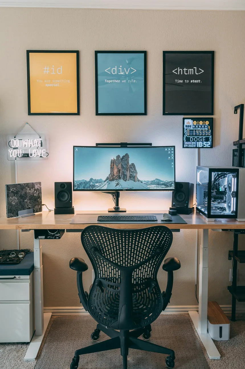 A tidy studio desk with an ultrawide monitor, framed HTML posters on the wall and a black mesh chair