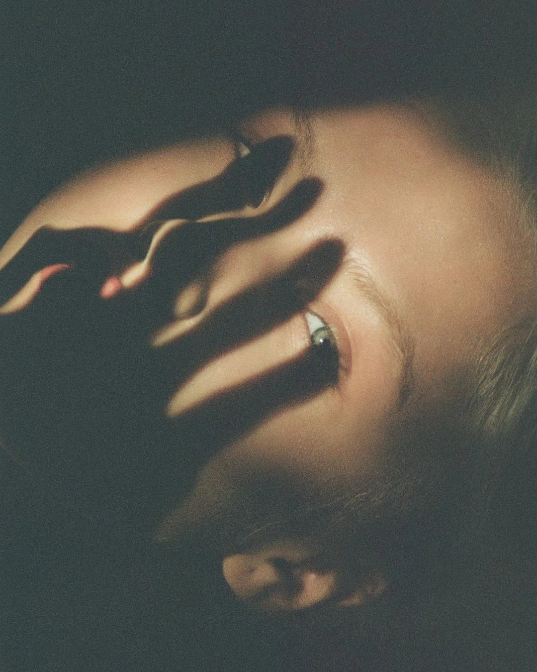 A grainy film frame: a hand wearing a wide silver ring resting across a face, lit from one side
